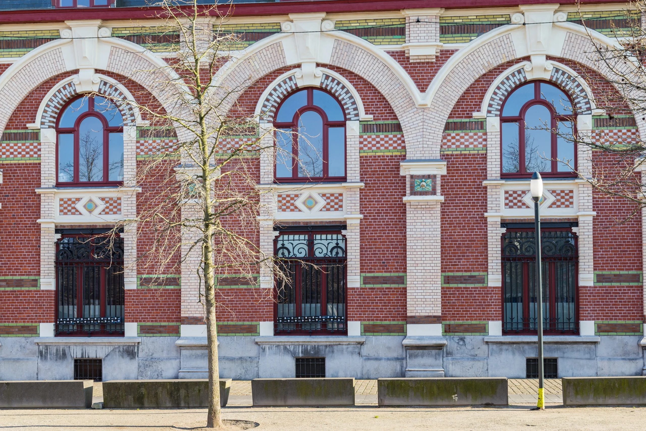 Façade en brique rouge ornée d’arcades de pierre claire, percée de hautes fenêtres bois cintrées peintes en bordeaux