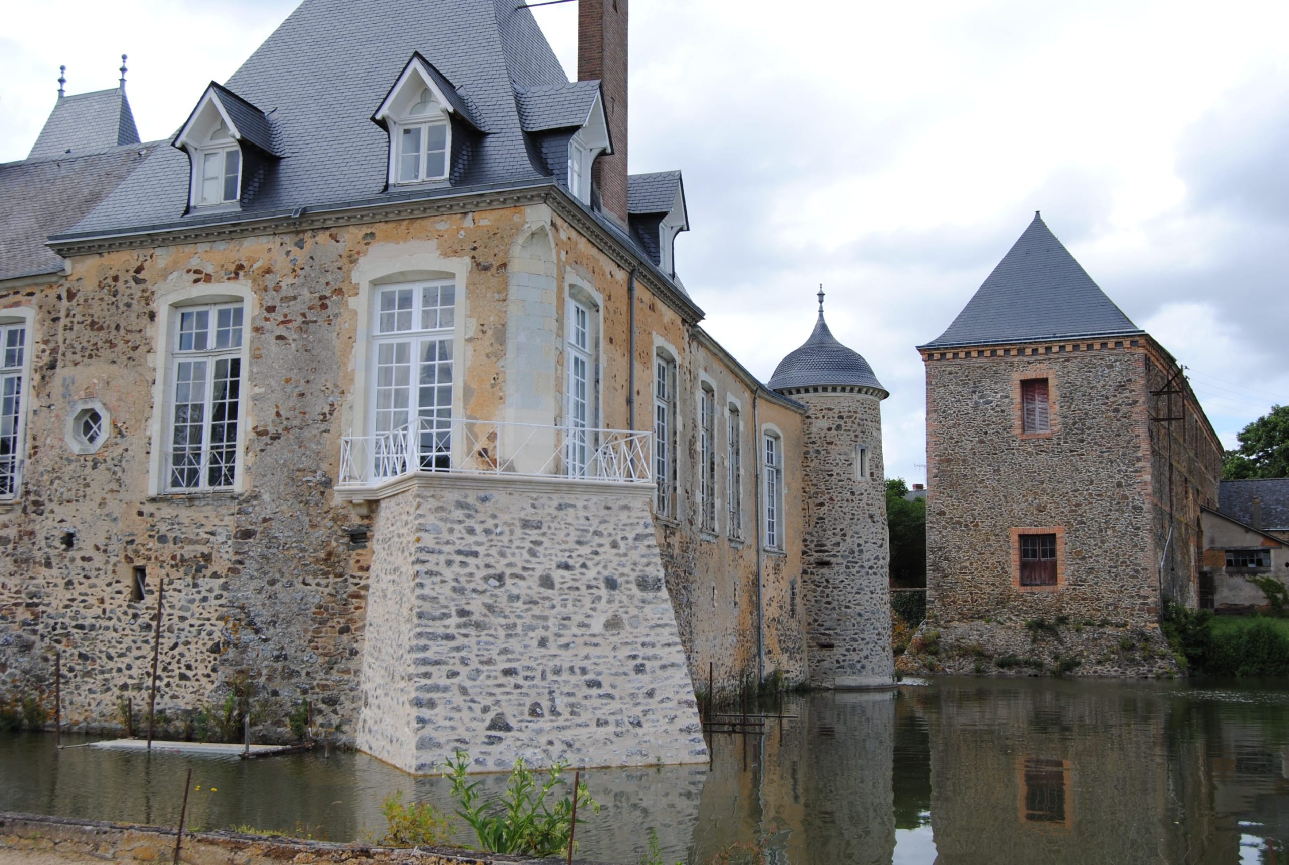 Château en moellons de pierre aux hautes fenêtres bois à petits bois, flanqué de tours et reflété dans des douves sous un ciel nuageux