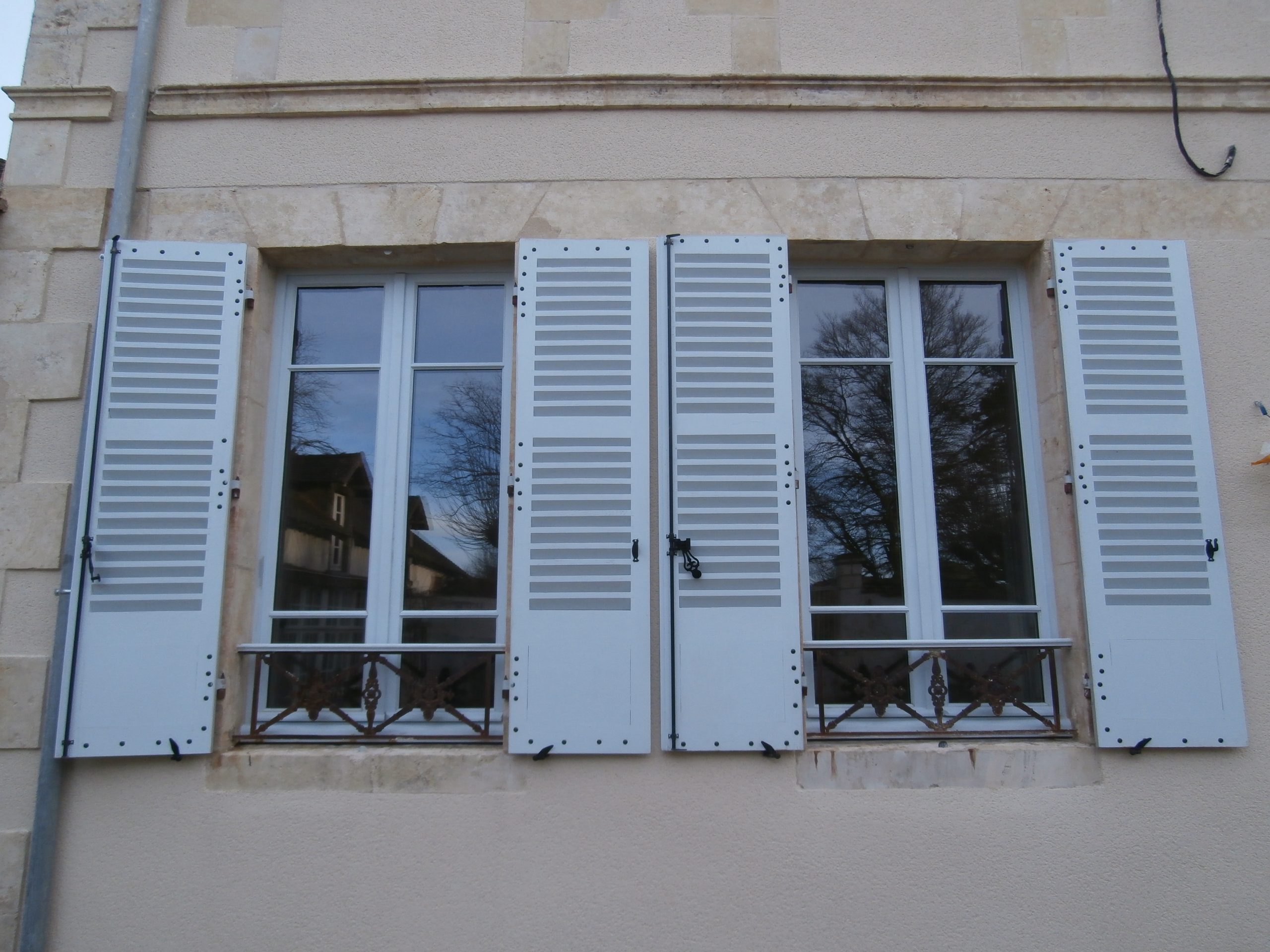 Deux fenêtres en bois à petits bois encadrées de persiennes grises ouvertes, sur une façade en pierre de taille avec appuis saillants